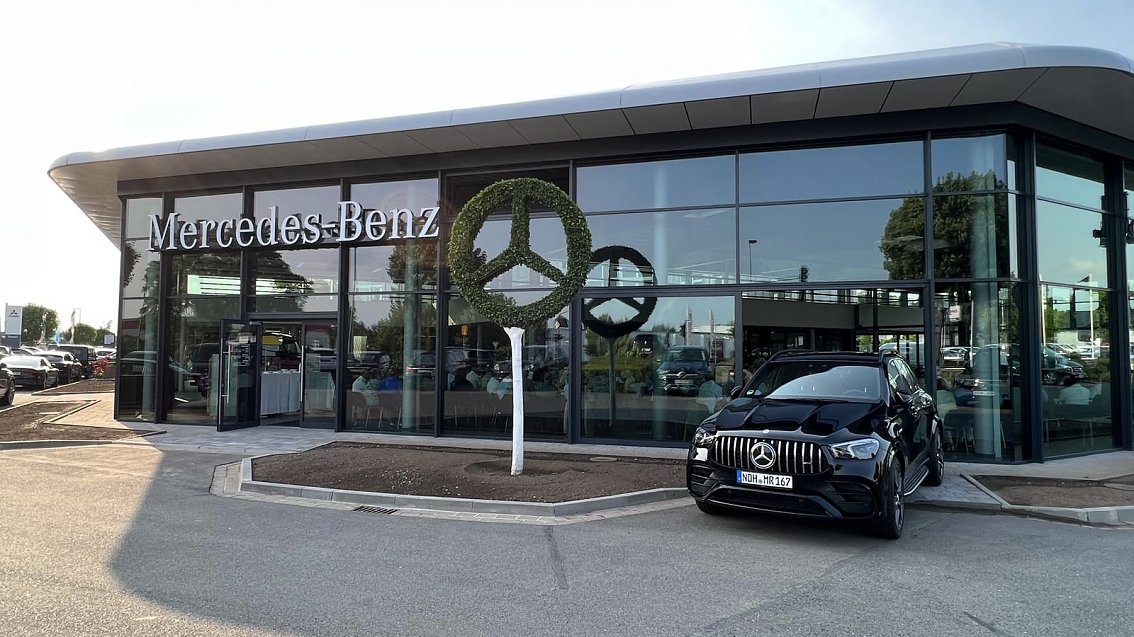 Mercedes-Benz Autohaus mit moderner Glasfassade, gro&szlig;em Mercedes-Stern aus Pflanzen vor dem Eingang und einem schwarzen Mercedes-AMG Fahrzeug davor.