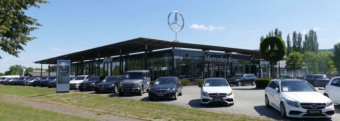 Mercedes-Benz Autohaus in Northeim mit gro&szlig;er Glasfassade, vielen ausgestellten Fahrzeugen auf dem Parkplatz, darunter SUVs und Limousinen. &Uuml;ber dem Geb&auml;ude ein gro&szlig;er Mercedes-Stern auf einem Mast, daneben eine gr&uuml;ne Heckenskulptur in Form des Mercedes-Sterns. Blauer Himmel, sonniges Wetter.