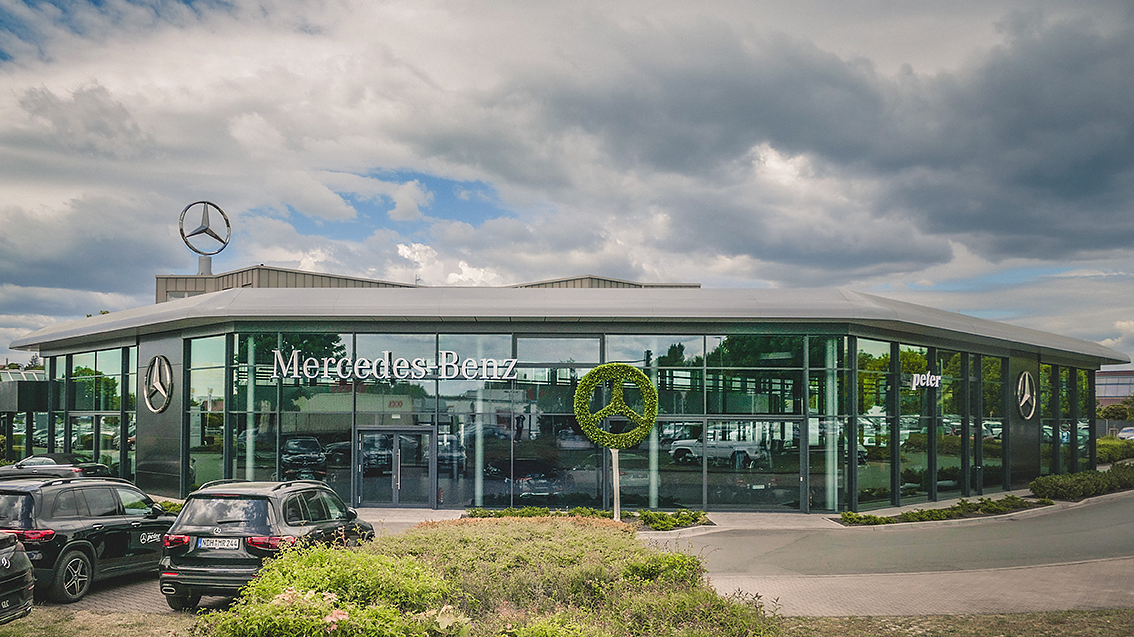Au&szlig;enansicht eines modernen Mercedes-Benz Autohauses Peter in Nordhausen mit gro&szlig;fl&auml;chiger Glasfassade, Mercedes-Benz Logos auf dem Dach und als Heckenschnitt, mehrere schwarze Fahrzeuge im Vordergrund, bew&ouml;lkter Himmel.