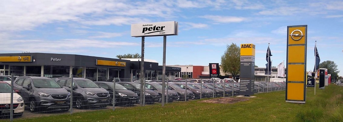 ADAC Standort in S&ouml;mmerda, Frohndorfer Stra&szlig;e 82. Im Vordergrund viele Opel-Fahrzeuge auf dem Hof hinter einem Zaun, links das Autohausgeb&auml;ude mit Opel- und Peter-Logos, daneben ein gro&szlig;er gelber ADAC-Pylon sowie weitere Fahnen und Schilder. Blauer Himmel mit Wolken.