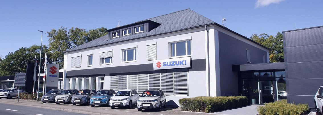 Suzuki-Autohaus in Nordhausen. Zweist&ouml;ckiges, graues Geb&auml;ude mit Suzuki-Logo und Schriftzug an der Fassade. Davor stehen mehrere Suzuki-Fahrzeuge in einer Reihe, links ein gro&szlig;es Suzuki-Schild. Umgeben von B&auml;umen, blauer Himmel mit wenigen Wolken.