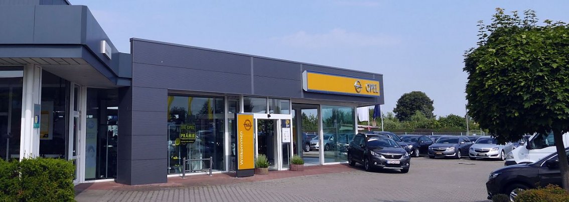 Au&szlig;enansicht des Opel-Autohauses der Autohaus Peter Gruppe in S&ouml;mmerda. Das Geb&auml;ude ist modern gestaltet, mit gro&szlig;en Glasfronten und einem auff&auml;lligen gelben Opel-Logo &uuml;ber dem Eingang. Mehrere Autos sind auf dem Parkplatz vor dem Autohaus zu sehen, umgeben von B&auml;umen und einer gepflegten Umgebung.