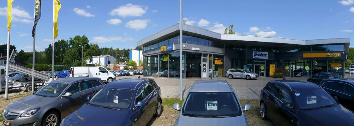 Au&szlig;enansicht des Opel-Autohauses in Osterode. Vor dem modernen, verglasten Geb&auml;ude mit markantem Dach sind zahlreiche Fahrzeuge geparkt, darunter verschiedene Opel-Modelle. Links wehen gelbe und schwarze Opel-Flaggen. Der Himmel ist blau mit ein paar Wolken, was dem Bild eine freundliche Atmosph&auml;re verleiht.
