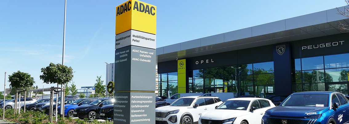 Au&szlig;enansicht des Standortes in Erfurt bei blauem Himmel. Links im Vordergrund ein gro&szlig;er gelb-grauer ADAC-S&auml;ulenpylon mit verschiedenen Serviceleistungen, dahinter eine Reihe geparkter Fahrzeuge. Dahinter das moderne Autohausgeb&auml;ude mit gro&szlig;en Glasfassaden und deutlich sichtbaren Markenlogos von Opel und Peugeot.