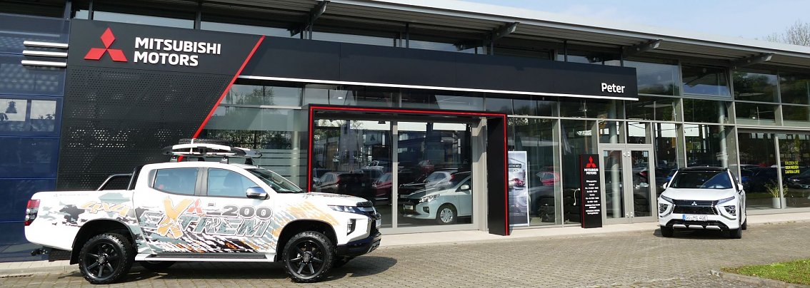 Eingang des Autohauses Peter in G&ouml;ttingen mit Mitsubishi-Motors-Logo, davor zwei Mitsubishi-Modelle, darunter ein auff&auml;llig gestalteter Pick-up.