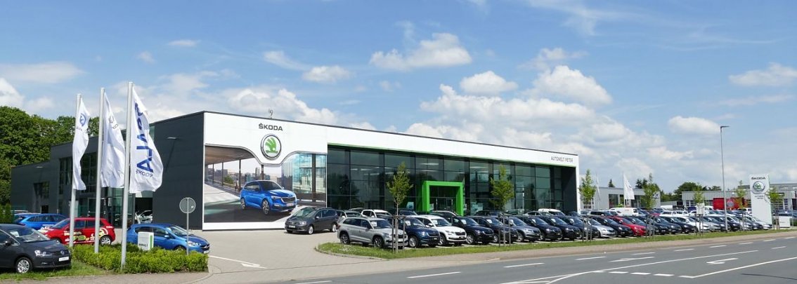 Moderne �koda-Autowelt Peter in Nordhausen mit gro&szlig;er Glasfassade und gr&uuml;nem Eingangsrahmen, davor viele geparkte Autos und wehende Fahnen bei blauem Himmel mit Wolken.