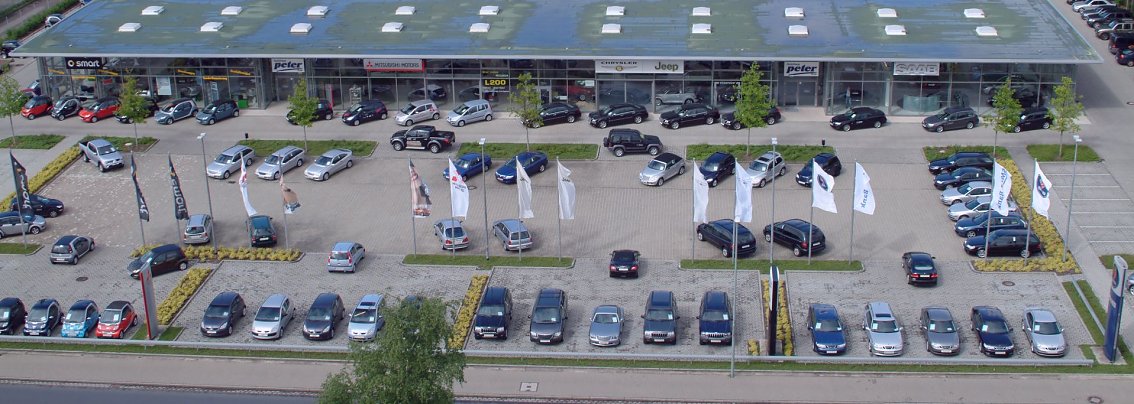 Luftaufnahme des ersten Mehrmarkenhauses der Autohaus Peter GmbH in G&ouml;ttingen, er&ouml;ffnet 2003. Vor dem gro&szlig;en, modernen Ausstellungsgeb&auml;ude mit mehreren Marken-Logos stehen zahlreiche Autos auf dem Parkplatz, umgeben von wei&szlig;en Flaggen und gepflegten Gr&uuml;nfl&auml;chen.