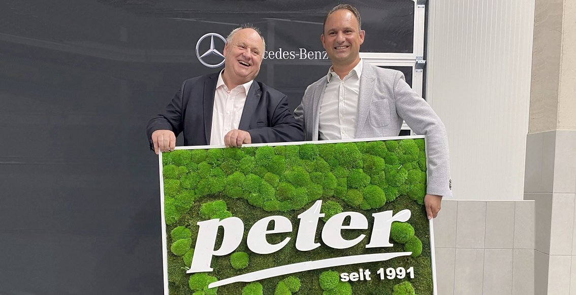 Helmut und Andreas Peter stehen nebeneinander und l&auml;cheln, im Hintergrund ein Mercedes-Benz-Logo. Sie halten ein gro&szlig;es Bild in die Kamera mit dem Schriftzug: Peter - seit 1991 (wei&szlig;e Schrift auf gr&uuml;nem Hintergrund aus Moos).