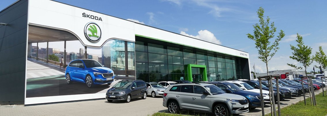 Modernes, im Jahre 2013 er&ouml;ffnetes, Skoda-Autohaus nach h&ouml;chstem europ&auml;ischen Standard mit gro&szlig;er Glasfassade, markantem gr&uuml;nem Eingangselement und auff&auml;lliger Fassadenwerbung mit einem blauen Skoda-SUV; davor zahlreiche ausgestellte Skoda-Modelle auf dem Vorplatz.