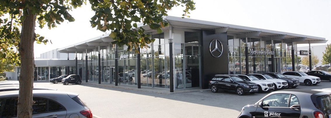 Modernes Mercedes-Benz-Autohaus der Peter Autozentrum Anhalt GmbH  in Dessau-Ro&szlig;lau mit gro&szlig;er Glasfassade und prominentem Mercedes-Stern, mehrere neue Fahrzeuge in Reih und Glied auf dem Au&szlig;engel&auml;nde; im Vordergrund B&auml;ume und geparkte Autos.
