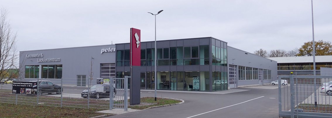 Modernes, gro&szlig;es Geb&auml;ude des Karosserie- und Lackierzentrums in Heilbad Heiligenstadt mit Glasfassade, grauer Fassade und rot-wei&szlig;em Logo; davor Parkpl&auml;tze und ein umz&auml;untes Gel&auml;nde.