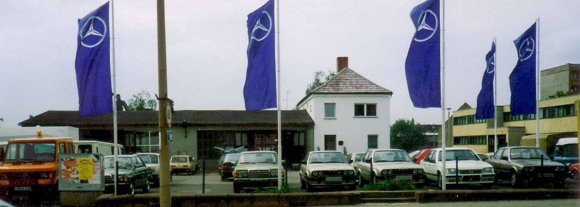 Historische Aufnahme des ersten Standorts der Autohaus-Peter-Gruppe in der Nordh&auml;user Zorgestra&szlig;e im Jahr 1990. Mehrere Mercedes-Benz Fahrzeuge sind vor einem Werkstatt- und B&uuml;rogeb&auml;ude geparkt, davor wehen 5 blaue Mercedes-Benz-Fahnen.