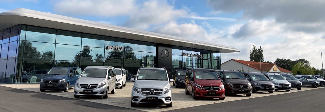 Mercedes-Autohaus Peter in Wittenberg mit moderner Glasfassade, davor eine Reihe von Mercedes-Vans in verschiedenen Farben bei bew&ouml;lktem Himmel.