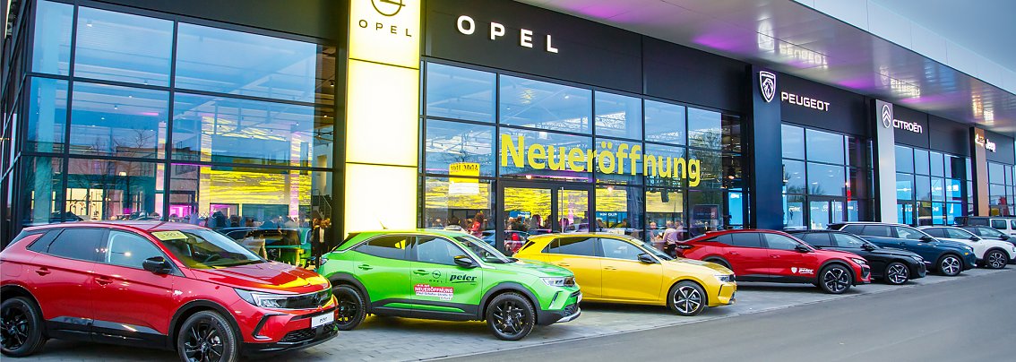 Mehrere farbige Autos stehen vor einer gro&szlig;en, modern beleuchteten Glasfassade des neugebauten STELLANTIS-Autohauses in Erfurt. Die Marken Opel, Peugeot und Citro&euml;n sind zu erkennen. Auf der Glasfassade steht in gro&szlig;en gelben Buchstaben das Wort Neuer&ouml;ffnung.