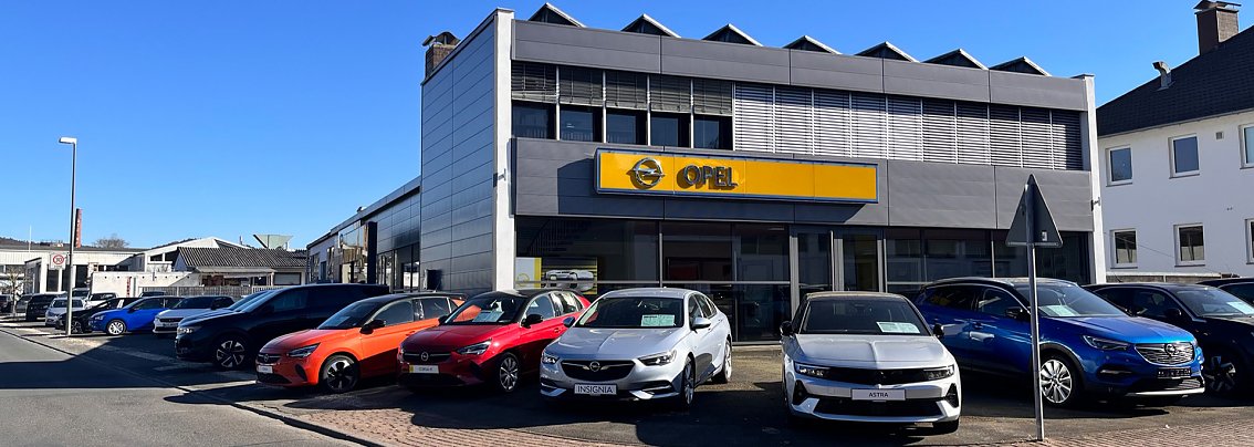 Au&szlig;enansicht des Opel-Autohauses in Witzenhausen mit mehreren bunten Fahrzeugen (u.a. rot, orange, blau, silber) auf dem Parkplatz vor dem Geb&auml;ude. &Uuml;ber dem Eingang ist ein gro&szlig;es gelbes Opel-Logo zu sehen. Der Himmel ist blau und die Sonne scheint.