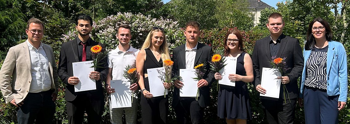 Gruppenfoto im Freien bei der feierlichen &Uuml;bergabe der Abschlusszeugnisse. Sechs junge Erwachsene mit Zeugnissen und Blumen, flankiert von zwei betreuenden Personen. Sonniger Tag, gr&uuml;ne Umgebung.