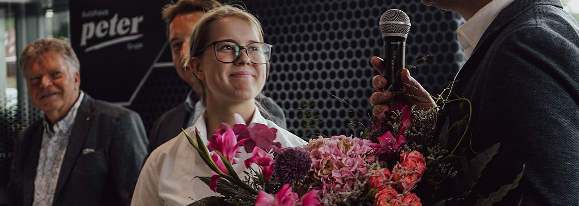 Lilly Hecker, frisch ausgebildete Automobilkauffrau und Jahrgangsbeste, steht l&auml;chelnd mit einem Blumenstrau&szlig; neben Gesch&auml;ftsf&uuml;hrer Andreas Peter, der in ein Mikrofon spricht. Im Hintergrund sind weitere Personen und das Logo der Autohaus-Peter-Gruppe zu sehen.