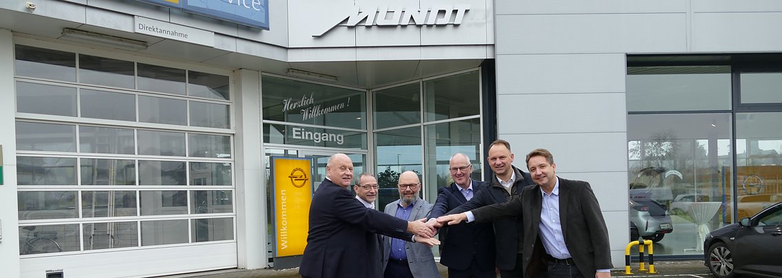 Mehrere Personen stehen vor einem Autohaus, halten die H&auml;nde partnerschaftlich &uuml;bereinander und l&auml;cheln in die Kamera