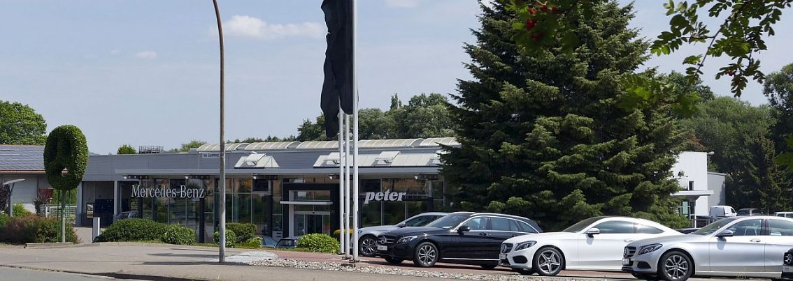 Mercedes-Benz Autohaus in Duderstadt, modern verglastes Geb&auml;ude mit Peter-Schriftzug, davor mehrere schwarze und wei&szlig;e Mercedes-Fahrzeuge aufgereiht, hohe Fahnenmasten mit schwarzen Fahnen, B&auml;ume und Str&auml;ucher im Hintergrund.