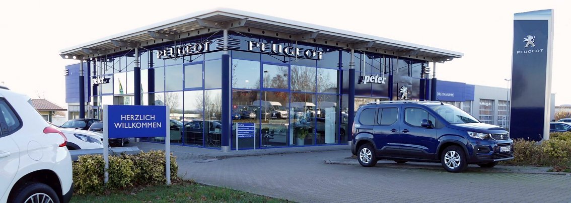 Peugeot-Autohaus in M&uuml;hlhausen mit moderner Glasfassade, gro&szlig;em Peugeot-Logo und einem blauen Willkommensschild im Vordergrund. Mehrere Fahrzeuge, darunter ein dunkelblauer Van, stehen vor dem Geb&auml;ude.