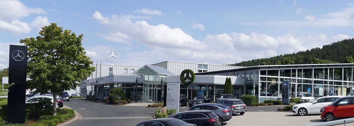 Au&szlig;enansicht des Mercedes-Benz Autohauses in Heiligenstadt bei sonnigem Wetter, mehrere geparkte Autos auf dem Parkplatz, gro&szlig;es Mercedes-Logo auf dem Dach und auf einer Stele, moderne Glasfassade und B&auml;ume im Hintergrund.