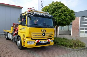 Gelber Abschlepp-LKW des Autohaus Peter im Auftrag des ADAC, Mercedes-Benz, mit der Aufschrift Autohaus Peter auf dem Dachschild und ADAC-Logo an der Front. Das Fahrzeug steht auf einem gepflasterten Platz neben einem Baum und einem grauen Geb&auml;ude mit rotem Dach.