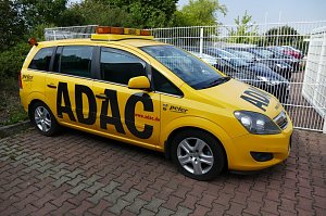 Gelber ADAC-Pannenhilfewagen vom Autohaus Peter, seitlich fotografiert auf einem gepflasterten Parkplatz, mit gro&szlig;em ADAC-Schriftzug und orangenem Warnlicht auf dem Dach. Im Hintergrund stehen weitere Fahrzeuge hinter einem wei&szlig;en Zaun.