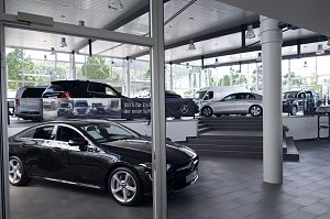 Innenraum eines Mercedes-Benz-Autohauses in Heiligenstadt. Im Vordergrund sieht man ein elegantes schwarzes Fahrzeug, im Hintergrund weitere ausgestellte Modelle auf verschiedenen Ebenen. Der Showroom wirkt modern, hell und gro&szlig;z&uuml;gig, mit gro&szlig;en Fensterfronten und viel Tageslicht.
