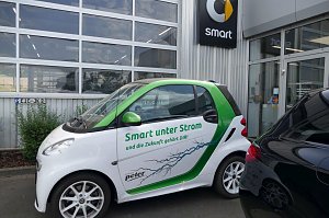 Wei&szlig;-gr&uuml;ner Smart mit der Aufschrift �Smart unter Strom und die Zukunft geh&ouml;rt DIR!�, geparkt vor einem Autohaus. Auf dem Auto ist ein Blitzmotiv abgebildet. Im Hintergrund ist das Smart-Logo an der Geb&auml;udewand zu sehen.