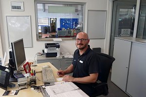 Freundlich l&auml;chelnder Mitarbeiter mit Brille und kurz&auml;rmeligem Poloshirt sitzt an einem Schreibtisch in einem Autohaus-B&uuml;ro. Vor ihm liegen Dokumente, ein Computer, Tastatur und B&uuml;romaterial. Im Hintergrund ist durch ein Fenster eine Werkstatt mit Reifenstapeln und Werkzeugen sichtbar.