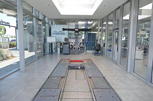 Helles, modernes Mercedes-Benz-Autohauses mit gro&szlig;en Glasfronten. In der Mitte eine Fahrzeughebeb&uuml;hne im Boden eingelassen, dahinter Werkstattausr&uuml;stung und ein Schreibtisch mit Stuhl. Rechts Glasb&uuml;ros mit Blick in den Innenraum.