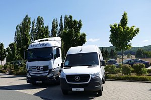 Zwei wei&szlig;e Mercedes-Benz Nutzfahrzeuge stehen nebeneinander auf einem Parkplatz. Links ein gro&szlig;er CharterWay-Lkw, rechts ein Transporter. Im Hintergrund gr&uuml;ne B&auml;ume, Autos und leicht h&uuml;gelige Landschaft unter blauem Himmel.