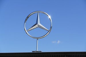 Gro&szlig;es silbernes Mercedes-Benz-Logo in Form des dreizackigen Sterns, montiert &uuml;ber einem Geb&auml;udedach. Strahlend blauer Himmel im Hintergrund, die Sonne spiegelt sich am Rand des Logos.
