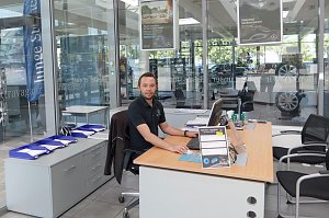 Mitarbeiter in schwarzem Mercedes-Benz Polohemd sitzt an einem modernen Holzschreibtisch im Autohaus, mit Computer, Dokumenten und Stühlen für Kunden, Glaswände mit Werbepostern und Fahrzeugen im Hintergrund.