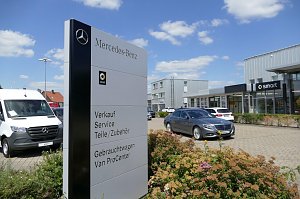 Schild mit Mercedes-Benz- und Smart-Logo vor einem modernen Autohaus mit Glasfassade. Auf dem Schild stehen Angebote wie Verkauf, Service und Gebrauchtwagen. Davor parken ein grauer Mercedes-Pkw und ein wei&szlig;er Mercedes-Van. Blumenbeet im Vordergrund, blauer Himmel mit wenigen Wolken im Hintergrund.