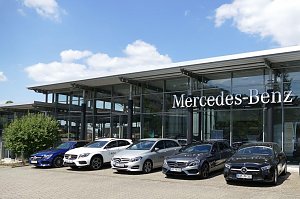 Mercedes-Benz Autohaus mit moderner Glasfassade und gro&szlig;em Mercedes-Benz-Schriftzug. Davor stehen f&uuml;nf Mercedes-Benz Fahrzeuge in verschiedenen Farben (blau, wei&szlig;, silber, grau, schwarz) nebeneinander aufgereiht. Blauer Himmel mit wei&szlig;en Wolken im Hintergrund.