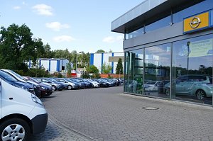 Außenansicht des Opel-Autohauses mit Glasfassade. Rechts ist das gelbe Opel-Logo über dem Eingang sichtbar. Vor dem Gebäude sind viele Autos geparkt, darunter Transporter und verschiedene Pkw-Modelle in einer Reihe. Im Hintergrund sieht man Bäume und Industriegebäude. Der Himmel ist blau mit einigen Wolken.