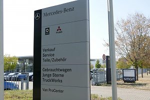 Großes Schild vor dem Mercedes-Benz Autohaus Peter in Dessau. Auf dem Schild stehen die Bereiche: Verkauf, Service, Teile/Zubehör, Gebrauchtwagen, Junge Sterne, TruckWorks und Van ProCenter. Oben Logos von Mercedes-Benz, Smart und Fuso. Im Hintergrund Glasfassade des Autohauses, parkende Autos und ein Baum.