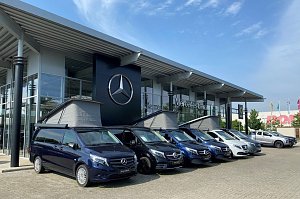 Außenansicht des Mercedes-Benz Autohauses in Dessau. Vor dem modernen Glasgebäude mit großem Mercedes-Stern-Logo stehen mehrere blaue und weiße Mercedes-Vans in einer Reihe. Drei Vans haben ausgeklappte Aufstelldächer.
