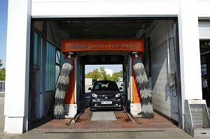 Ein grauer Smart fährt gerade in eine Autowaschanlage mit orangefarbener Beschriftung �Christ CAR WASH�. Links und rechts hängen graue Bürsten.
