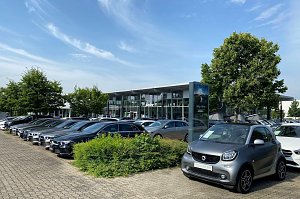 Außenansicht des Mercedes-Benz Autohauses Peter in Dessau. Im Vordergrund steht ein grauer Smart mit dem Schild �Gebrauchtwagen�. Daneben viele schwarze und silberne Mercedes-Benz Fahrzeuge in einer Reihe auf dem Parkplatz. Im Hintergrund modernes Glasgebäude mit großen Fenstern, Bäumen und blauem Himmel mit leichten Wolken.