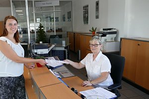 Eine junge Frau überreicht einer Mitarbeiterin an einem Empfangsschalter freundlich ein Dokument. Die Mitarbeiterin sitzt hinter dem Schreibtisch und trägt eine Brille. Beide Frauen lächeln. Im Hintergrund sieht man Büroräume und Schränke.