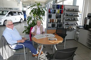 Zwei &auml;ltere Kunden sitzen im Wartebereich eines Mercedes-Autohauses an einem Tisch mit Zeitungen, im Hintergrund ein wei&szlig;er Van, Prospektst&auml;nder und Farbmusterwand.