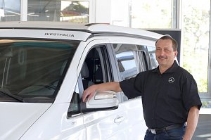 Ein Autohaus-Mitarbeiter im schwarzen Hemd mit wei&szlig;em Mercedes-Logo steht l&auml;chelnd neben einem wei&szlig;en Westfalia-Campervan, lehnt mit einer Hand an der Fahrert&uuml;r.