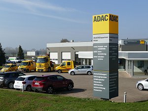 ADAC-Standort in Nordhausen; im Vordergrund eine gro&szlig;e gelb-graue ADAC-Stele mit Beschriftung Mobilit&auml;tspartner Peter. Dahinter mehrere gelbe ADAC-Pannen- und Abschleppwagen und ein modernes, graues Geb&auml;ude mit Glasfassade und ADAC-Logo. Im Vordergrund parkende Autos, im Hintergrund Felder und B&auml;ume bei klarem Himmel.