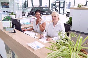 Zwei Mitarbeiterinnen an der Rezeption des �koda-Autohauses, eine telefoniert, die andere schreibt. Helle, moderne Umgebung mit Pflanzen und Autos im Hintergrund.