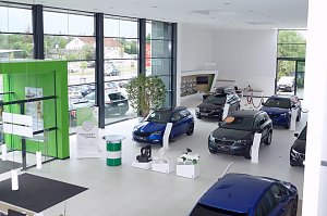 Heller �koda-Showroom mit großen Fensterfronten, mehreren ausgestellten Fahrzeugen, einem markanten grünen Eingangselement und Zubehör auf weißen Podesten.