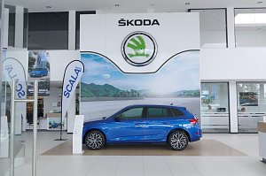 Innenansicht des �koda-Autohauses mit großem weißen �koda-Logo an der Wand. In der Mitte steht ein blauer �koda Scala, flankiert von Werbefahnen und Postern.