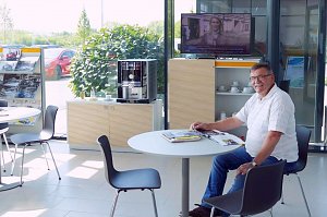 Ein älterer Mann sitzt lächelnd an einem runden Tisch in einer hellen Lounge im Opel-Autohaus Peter in Nordhausen. Auf dem Tisch liegen Zeitschriften. Im Hintergrund stehen ein Kaffeeautomat, ein TV mit Nachrichten und Prospektständer. Durch die großen Fenster sieht man Autos und grüne Pflanzen draußen.