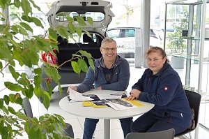 Ein Mann und eine Frau sitzen an einem Tisch im Autohaus, mit Broschüren und Prospekten vor sich, im Hintergrund ein geöffneter Kofferraum eines Autos und helle Glasfronten.